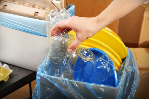 A close-up image of a person's hand gripping a clear plastic bottle above a collection of recycling containers. The containers are made of durable plastic with a smooth finish, and they are stored inside a transparent blue recycling bag. The yellow and blue bins, used for separating waste types, are partially obscured but clearly identifiable by their vivid colours. The background shows a tidy indoor environment with a grey surface and a wooden surface, possibly part of a storage or sorting area. The scene demonstrates typical waste sorting activities relevant to rubbish removal services, with an emphasis on recycling practices. This visual supports local rubbish and waste management efforts in the Hornsey area, aligned with Garden Clearance Hornsey's focus on professional waste collection and environmental responsibility.