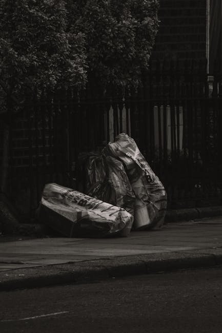 A black wheeled rubbish bin with the label 'ST. JOHN'S' positioned on a sidewalk at night, with its lid open revealing various discarded paper and cardboard packaging on top, some skewed and leaning a