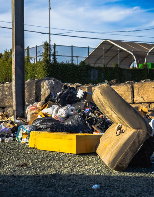 A large collection of mixed household waste and rubbish is piled up around several overflowing rubbish bins on a paved urban street. The rubbish includes cardboard boxes, plastic bags filled with tras