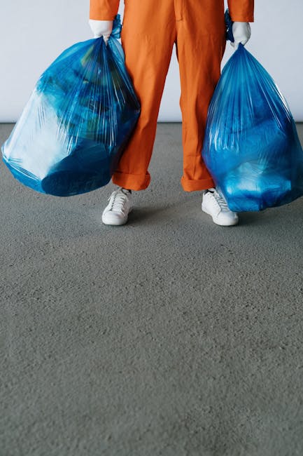 A person wearing orange work trousers and white sneakers is standing on a grey concrete floor, holding two large blue plastic rubbish bags filled with waste materials. The individual's hands are glove