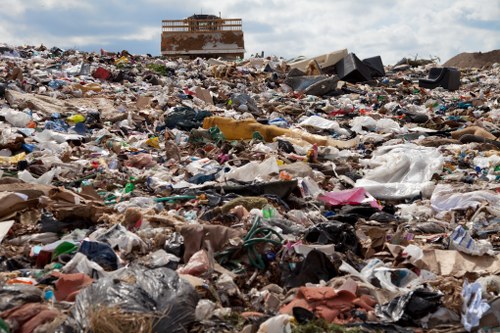 A vast open landfill site covered with a large accumulation of mixed waste materials, including plastic bags, cardboard, paper, and discarded clothing, spread across the ground in front of a partially constructed or abandoned building structure in the background. The scene shows a densely packed area of refuse extending towards the horizon, with some piles of rubbish visibly stacked or spread unevenly. The sky above appears cloudy, and the overall environment suggests an area designated for waste disposal or processing, typical of rubbish removal sites. This image illustrates the importance of professional waste management services, such as those offered by Garden Clearance Hornsey, to assist with the removal and proper disposal of household or garden rubbish within London and the surrounding areas.